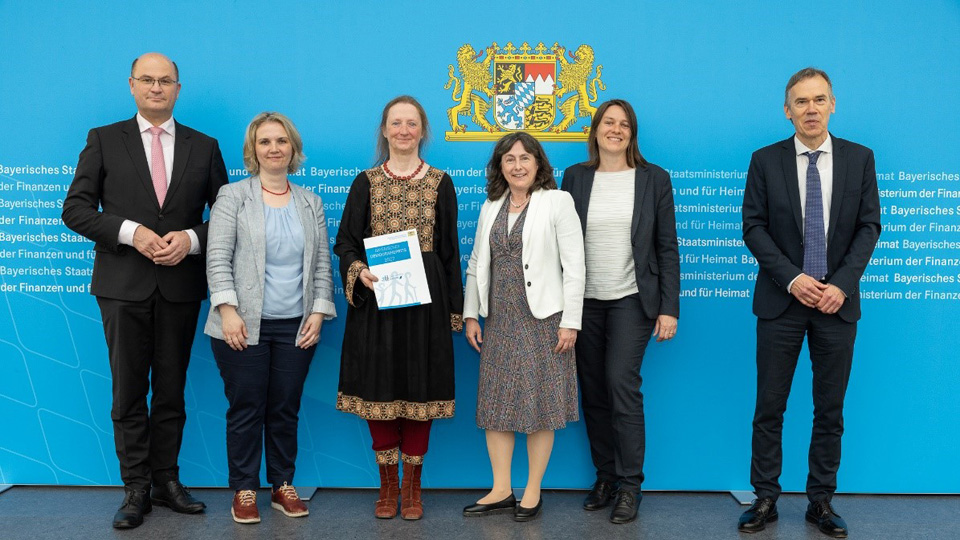 Eingerahmt von zwei Männern stehen vier Frauen vor dem Bayerischen Landeswappen