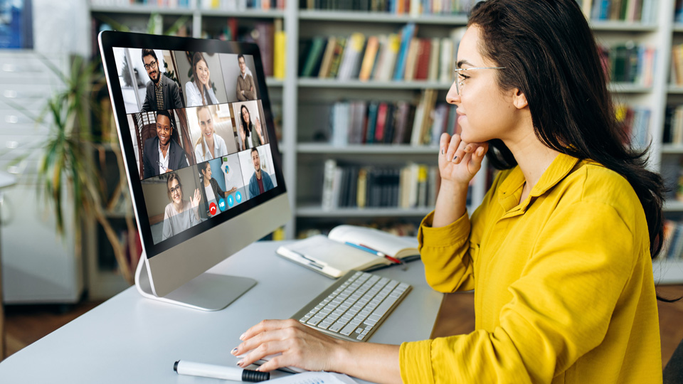 Eine Frau sitzt an einem Schreibtisch und schaut in einen Monitor mit mehreren Videokonferenzteilnehmenden.