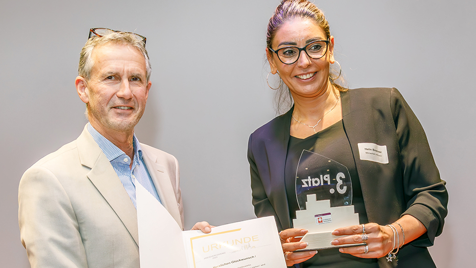 Ein Mann und eine Frau stehen nebeneinander und präsentieren eine Urkunde und eine Pokal.