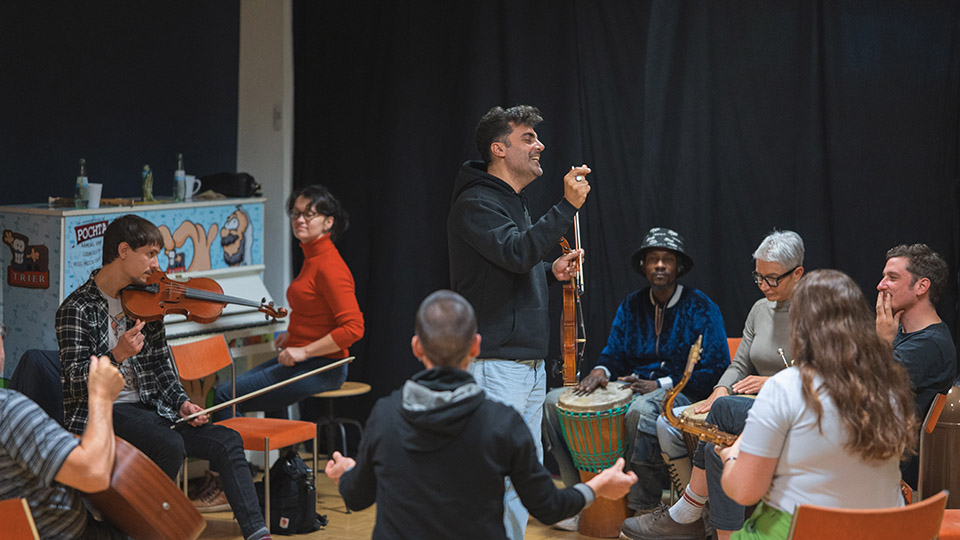 Viele Personen sitzen im Kreis und spielen ein Instrument. Ein Mann steht in deren Kreismitte.