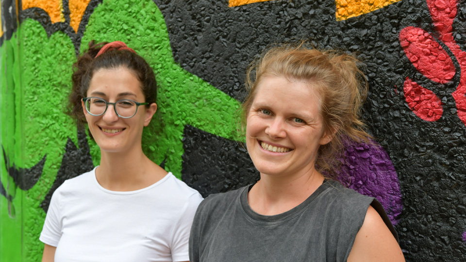 Zwei Frauen stehen vor einer mit Graffiti besprühten Wand und lächeln in die Kamera.